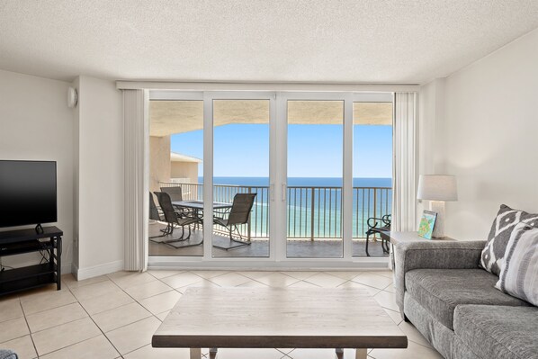A living room with a TV, a sofa, a coffee table, and a balcony with a view of the ocean.