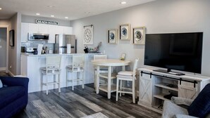 A modern kitchen with white cabinetry, a microwave, and a refrigerator. There is a dining area with a white table and chairs, and a TV mounted on a wooden stand.