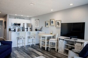 A modern kitchen with white cabinetry, a microwave, and a refrigerator. There is a dining area with a white table and chairs, and a TV mounted on a wooden stand.