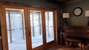 A room with a wooden piano, a clock on the wall, and a large window with a view of snow-covered trees.