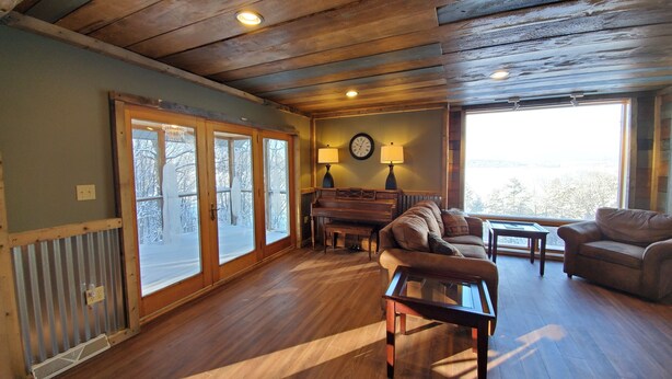 A spacious living room with wooden flooring, a sofa, a coffee table, a piano, and a large window offering a view of the outdoors.