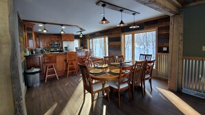 A rustic cabin interior with a wooden dining table, chairs, and a kitchen area with wooden cabinets and a large window.