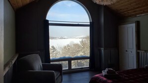 A room with a large arched window, a bed, a chair, and a wooden ceiling.