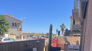 Une terrasse extérieure avec des meubles en osier, un mur de pierre orné d’une statue de lion, et une vue sur les voitures garées et les bâtiments en arrière-plan.