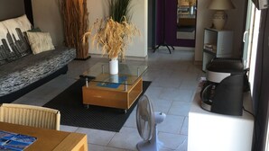 Un salon comprenant un canapé, une table basse et une table à manger. On trouve sur la table basse un ventilateur, une lampe et une plante.