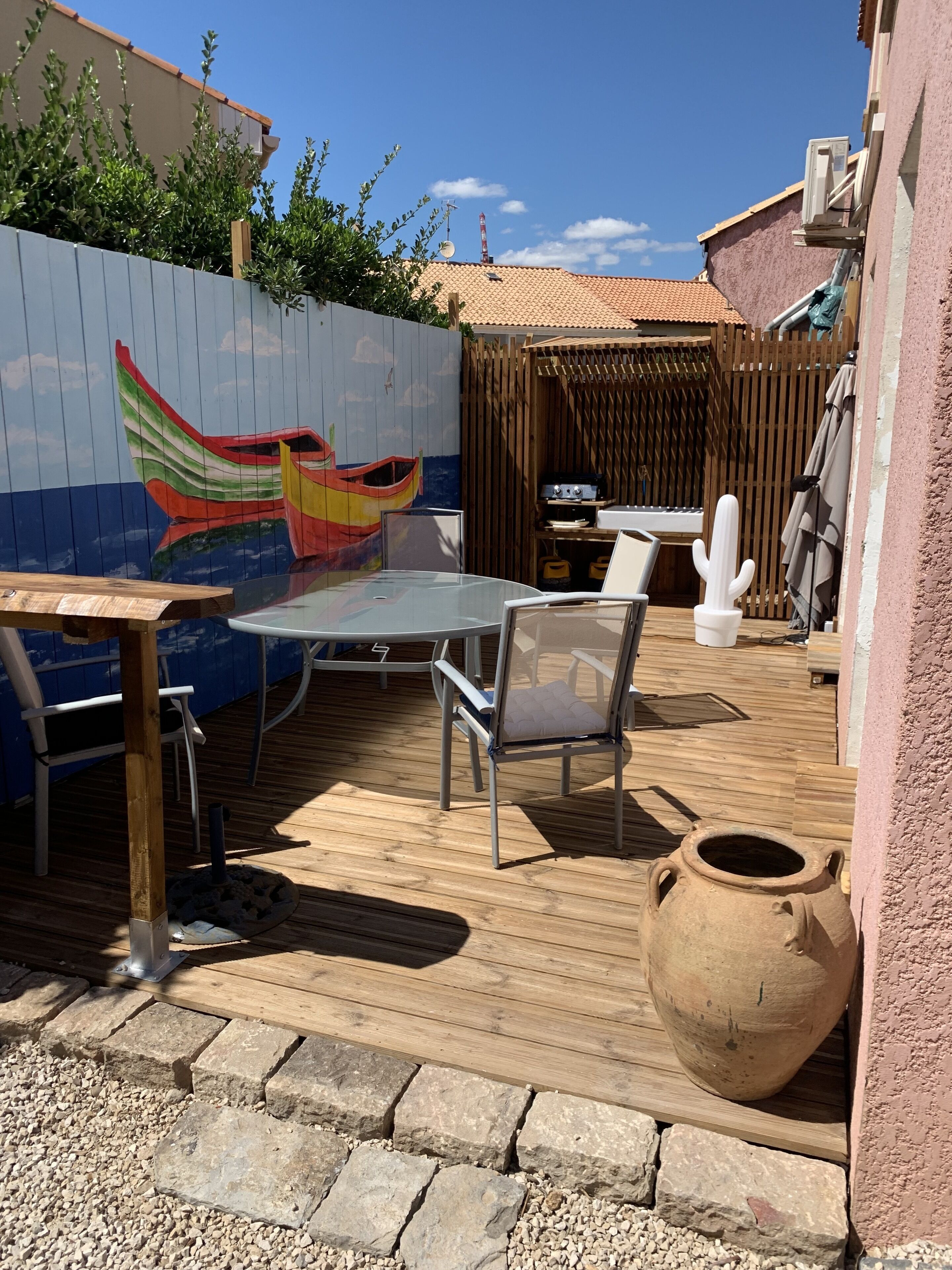 Une terrasse extérieure avec une plateforme en bois, une table en verre, des chaises en métal, un grand pot en argile et une fresque représentant un bateau sur le mur.