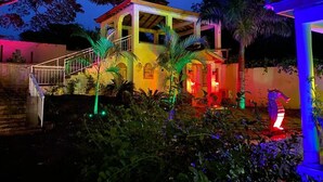 A two-story house with a covered porch, surrounded by lush greenery and illuminated by colorful lights. - Family Friendly~Chef Service- Book 8 to 14 Rooms (Sosua)