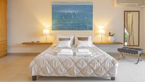 Un lit bien fait, recouvert d’une courtepointe matelassée, deux tables de chevet avec des lampes, un climatiseur fixé au mur et un tableau représentant des voiliers accroché au mur.