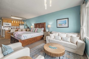A bedroom with a bed, bedside tables, a sofa, a round wooden table, and a kitchen area with cabinets and appliances.