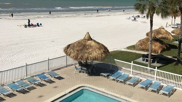 A pool area with lounge chairs and a thatched-roof structure, overlooking a beach with people and palm trees. -  Updated and Restored Beachfront Condo with Spectacular Views of the Gulf (Redington Shores)