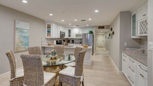 A modern kitchen with white cabinets, a glass dining table, wicker chairs, and a stainless steel refrigerator. -  Updated and Restored Beachfront Condo with Spectacular Views of the Gulf (Redington Shores)