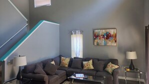 A living room with a grey sofa, a coffee table, a ceiling fan, a painting, and a staircase with teal accents. -  The Wilson Wilds, A Peaceful 4BR Family Retreat, Private Pool 15 Mins to Disney (Davenport)