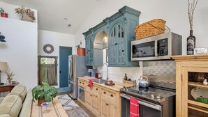 A compact kitchen with teal cabinets, a stainless steel refrigerator, a microwave, and a wooden cabinet with a basket on top.