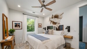 A bedroom with a large bed, a ceiling fan, a window with curtains, a wooden desk, and a wall-mounted shelf.