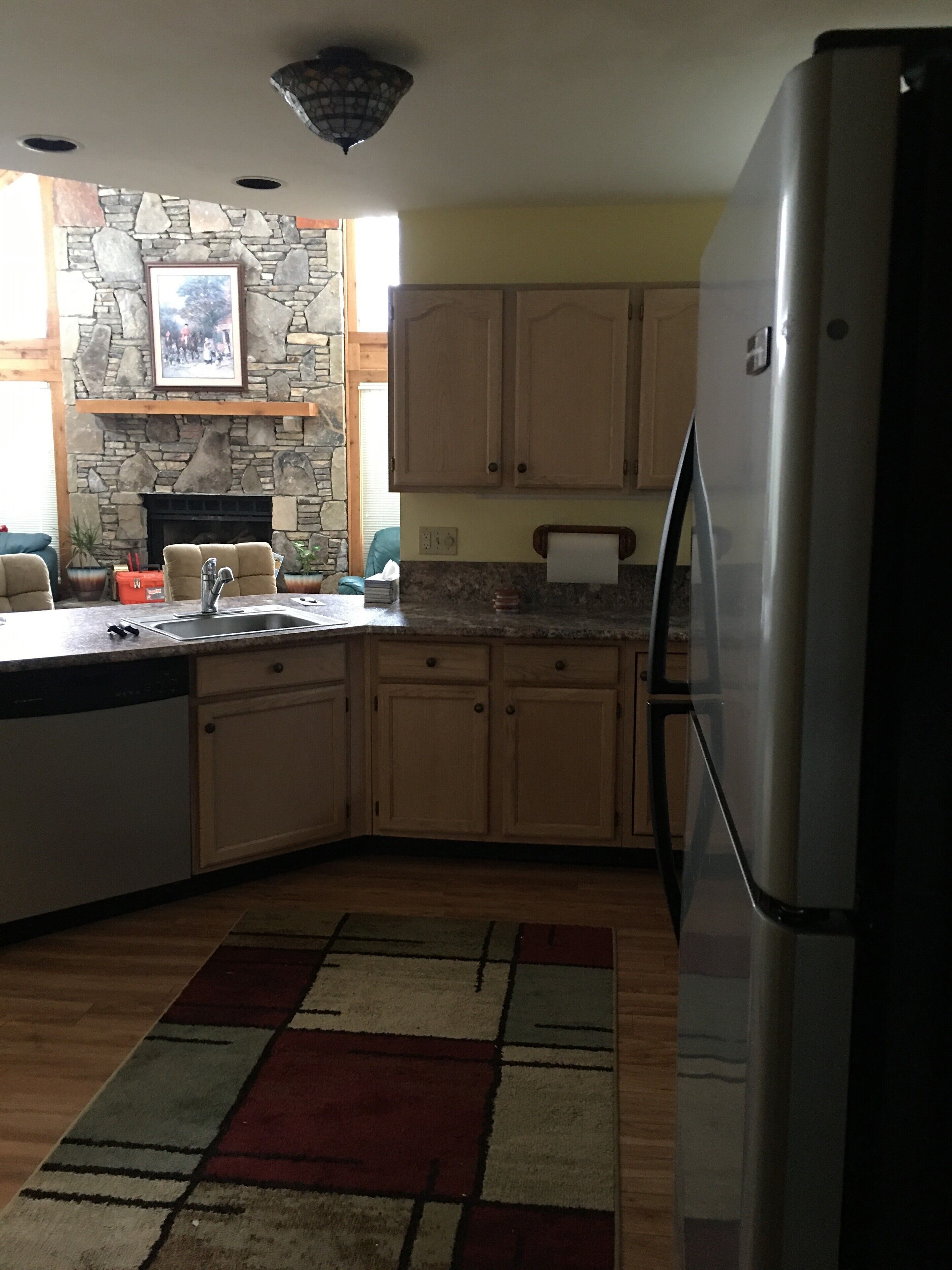 A kitchen with a stone fireplace, wooden cabinets, a stainless steel refrigerator, and a patterned rug.