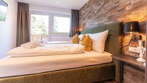 A neatly made bed with white linens and multiple pillows, a wooden accent wall, a bedside table with a lamp, and a window with curtains. - Tevini Boutique Suites - Apartment Water, balcony (Zell am See)