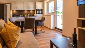A modern living room with a sofa, a dining table with chairs, a kitchen area, and a TV mounted on the wall. - Tevini Boutique Suites - Apartment Earth, balcony (Zell am See)