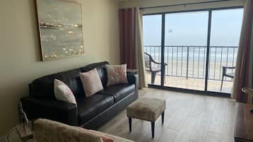 Una sala de estar con un sofá de cuero negro, un sillón estampado y un pequeño reposapiés. Se observa un ventilador de techo, y la habitación tiene un gran ventanal corredizo que da a un balcón con vista a la playa.