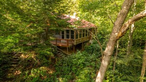 Una cabaña tipo casa en árbol rodeada de un bosque denso, con una terraza de madera y un tejado rojo.