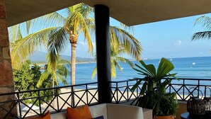 Ein Balkon mit einer weißen Couch, Kissen in Orange und Blau mit Muster, einem Blumentopf und einem Tisch mit Kerzen und einem Buch.