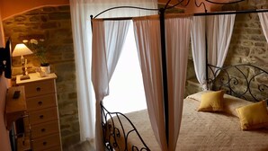 A bedroom with a stone wall, a four-poster bed, a nightstand with a lamp, and a dresser. - Fonte Rosa farmhouse with pool (Fiastra)