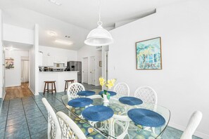 Een eetgedeelte met een glazen tafel, omringd door witte stoelen met blauwe kussens, een witte hanglamp en een schilderij aan de muur.