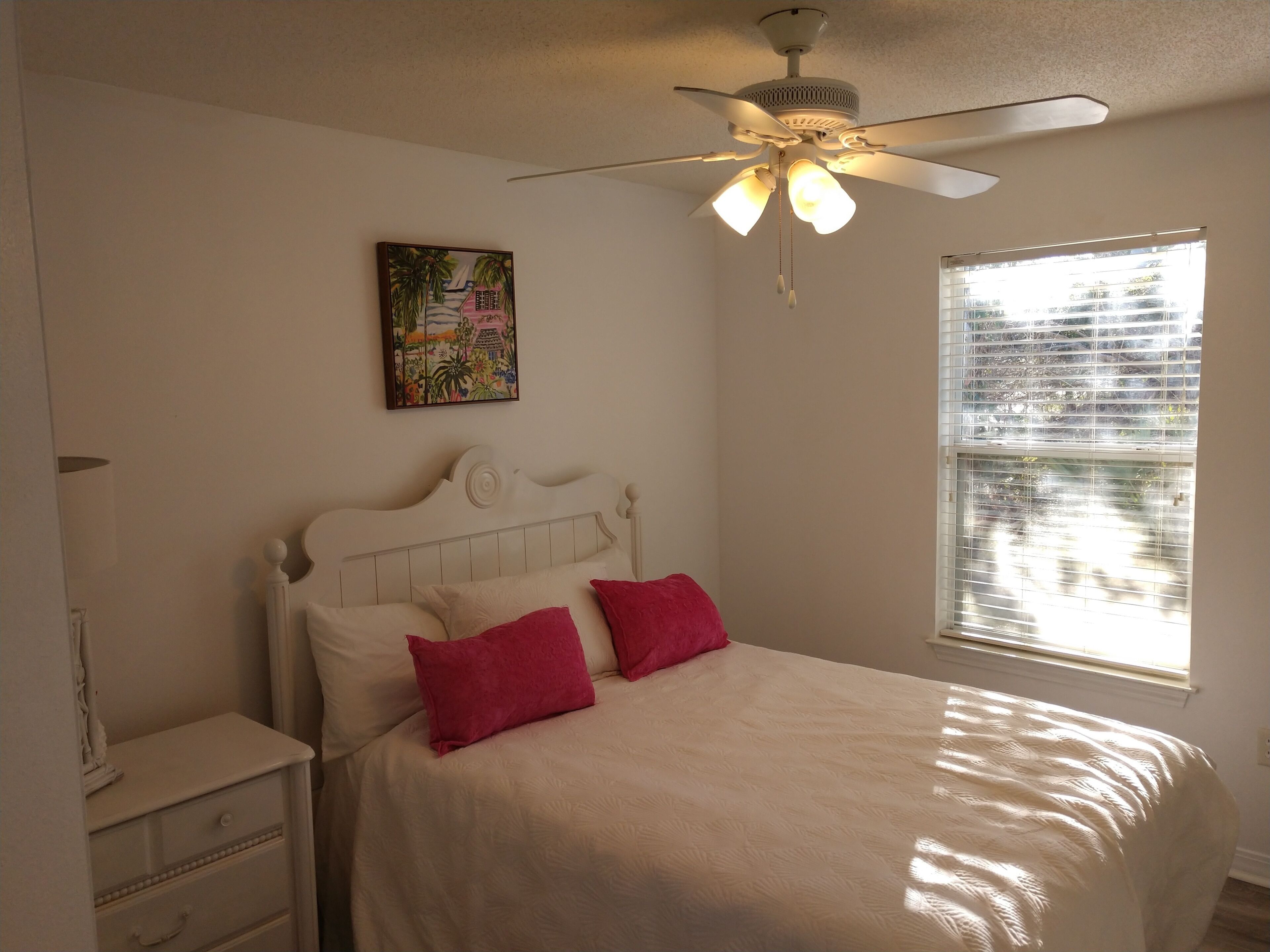 A bedroom with a bed, a ceiling fan, a window with blinds, and a painting on the wall.