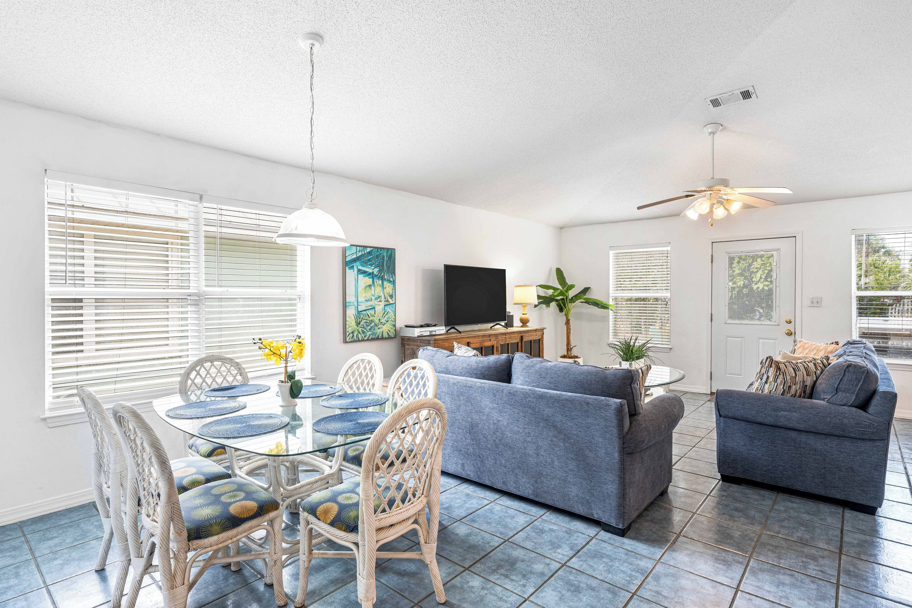 A spacious living room with a dining area, a gray sofa, wicker chairs, a TV, and a ceiling fan.