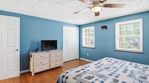 Une chambre à coucher comprenant un lit, une commode, un téléviseur, un ventilateur de plafond et deux fenêtres équipées de stores.