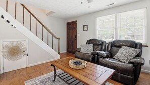 A living room with a wooden coffee table, a leather sofa with cushions, a staircase with wooden railings, and a door with a decorative glass panel.