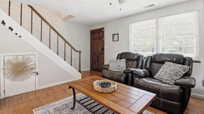 A living room with a wooden coffee table, a leather sofa with cushions, a staircase with wooden railings, and a door with a decorative glass panel.