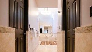 Un couloir menant à une salle de bain dotée d’un grand miroir, d’un lustre et d’un tapis à rayures.