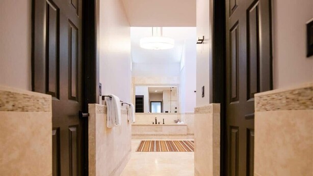 Un couloir menant à une salle de bain dotée d’un grand miroir, d’un lustre et d’un tapis à rayures.