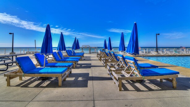 A beachside lounge area with rows of wooden lounge chairs and blue cushions, overlooking a clear blue sea. - Spacious Beachfront Condo Located on Grand Lagoon  (Panama City)