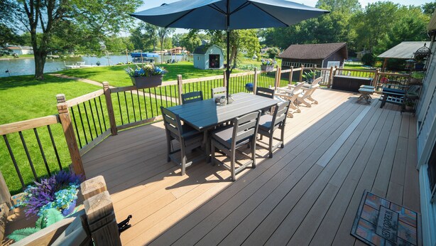 Une terrasse en bois avec une table et des chaises, un parasol bleu et une vue sur un lac et des bateaux.