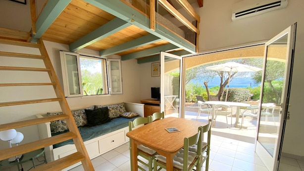 A cozy room with a wooden staircase, a sofa with cushions, a dining table with chairs, and a large sliding glass door leading to an outdoor patio with a view of the sea.