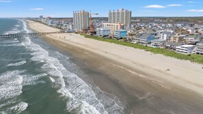 A coastal cityscape with a wide sandy beach, a pier extending into the ocean, and high-rise buildings in the background.