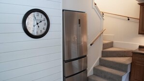 A kitchen with a stainless steel refrigerator, a wall-mounted clock, and a staircase with a handrail.