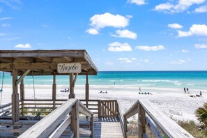 A wooden beach deck with a sign reading 'The Cabo' overlooking a sandy beach and clear blue ocean.