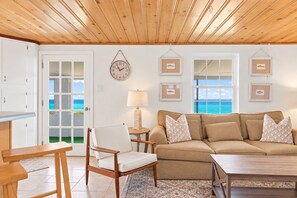 A living room with a wooden ceiling, a beige sofa, a wooden chair, a small table, and a view of the ocean through a door.