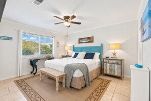 A bedroom with a bed, a ceiling fan, a nightstand with a lamp, a bench, and a window with blinds.