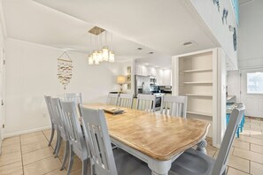 A dining area with a wooden table and grey chairs, a hanging light fixture, and a kitchen in the background.
