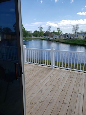 Eine Holzterrasse mit weißer Reling blickt auf ein ruhiges Gewässer, umgeben von Häusern und Bäumen.