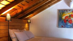 A bedroom with a sloped ceiling, wooden beams, a bed with white bedding, two bedside lamps, and a painting on the wall. - Nice country studio for 2-4p., shared pool, countryside&beach (20 km) (cercal do alentejo)