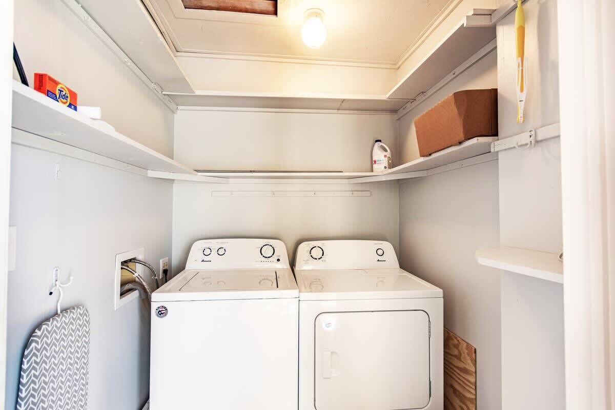 A laundry room with two white washing machines, a shelf with a box of Tide detergent, and a hanging iron board.