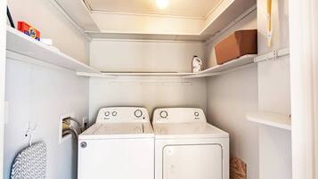 A laundry room with two white washing machines, a shelf with a box of Tide detergent, and a hanging iron board.