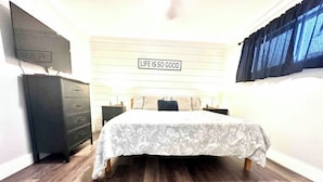 Une chambre à coucher avec un lit, une commode, un téléviseur et un panneau mural sur lequel on peut lire : « LA VIE EST BELLE ».