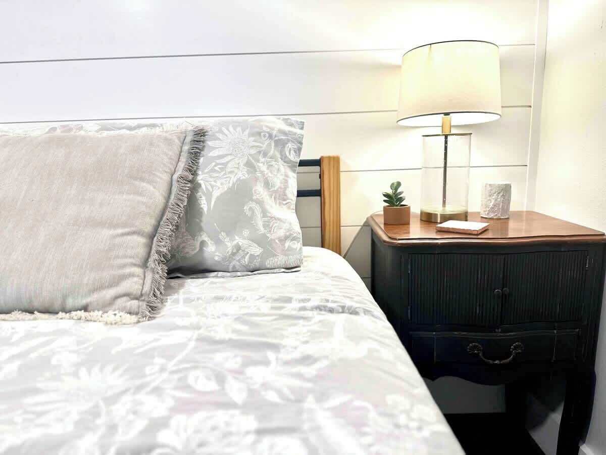 A neatly made bed with a white and gray patterned bedspread, a white pillow with a floral design, and a small bedside table with a lamp, a plant, and a few items.