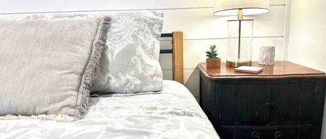 A neatly made bed with a white and gray patterned bedspread, a white pillow with a floral design, and a small bedside table with a lamp, a plant, and a few items.