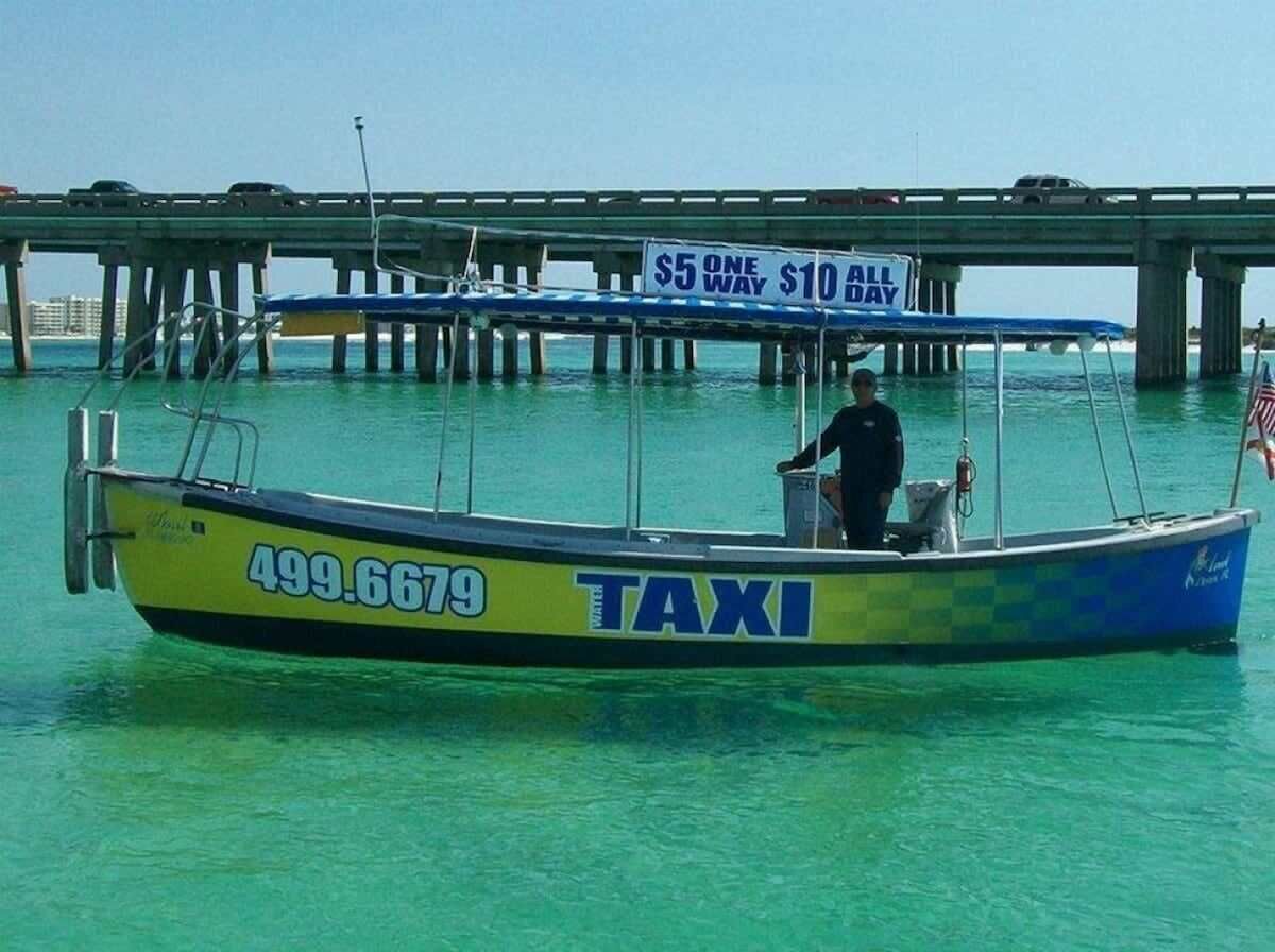 A boat with the word "TAXI" and the number 499.6679 on its side, docked near a bridge with a sign that reads "$5 ONE WAY $10 ALL DAY".
